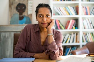 Emily Bernard faces the camera, seated at a table, with one hand touching her face. A wall of bookshelves and a painting of a Black woman are in soft focus behind her.