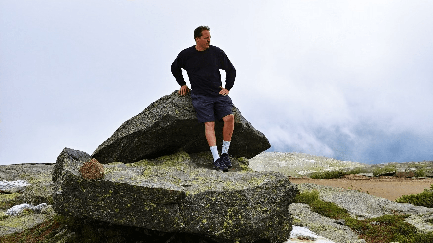 Malcom Greene oudoors, standing on a large rock, leaning against another large rock, looking to the left.